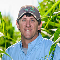 White man with brown hair and a brown hat stands in a corn field on a sunny day