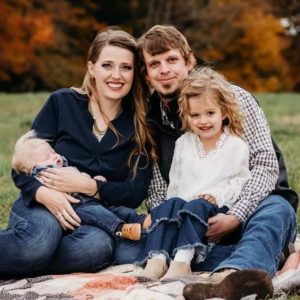 Amos and Tina Troester sitting for a family portrait on a blanket in the grass with two young children on their laps