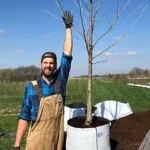 Zach Burhenn stands next to a potted tree, his arm stretched up to reach the upper branches, as he smiles for the camera. 