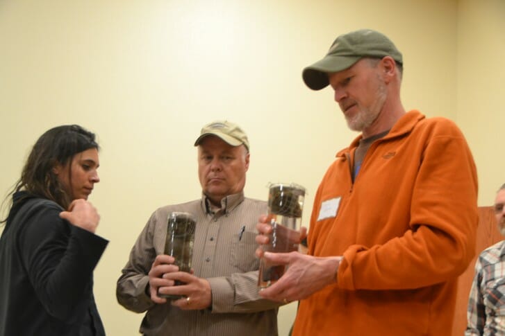 PFI member Clark Porter far right) and another attendee assist Tina Cibula, NRCS, conduct a "slake" test. The no-till soil Clark is holding keeps its form while the tilled soil in the other beaker breaks up rapidly when plunged into the water.